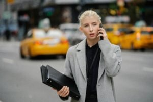 Focused young businesswoman in gray coat standing on busy street with folders and having conversation on mobile phone